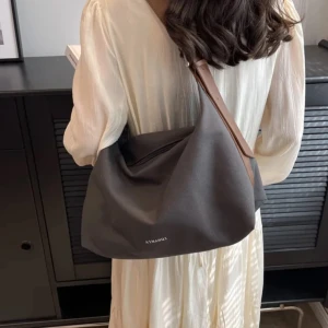 Grå axelväska från AIMANQI - Snygg grå axelväska från AIMANQI med justerbart brunt axelband i skinnimitation. Väskan är rymlig och har en enkel, stilren design med diskret logga framtill. Perfekt för dig som vill ha en praktisk och trendig väska till vardags.