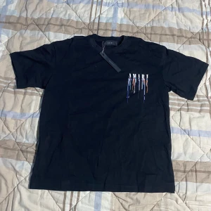 Svart AMIRI t-shirt med färgstänk - Svart t-shirt från AMIRI med coolt tryck av droppande färg och stor logga både fram och bak. T-shirten har rund hals, korta ärmar och är gjord i mjuk bomull. Perfekt för dig som vill sticka ut med streetstyle-vibe.
