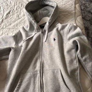 Grå hoodie från Polo Ralph Lauren - Snygg ljusgrå hoodie från Polo Ralph Lauren med dragkedja framtill, huva med snörning och klassisk broderad logga på bröstet. Mjuk bomullsblandning och två fickor framtill gör den både chill och stilren.