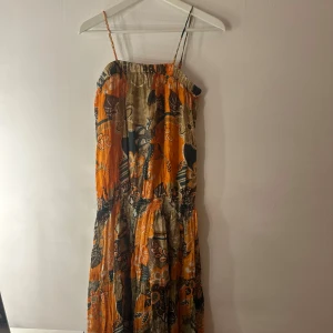 Blommig långklänning från etc. - Färgstark långklänning med smala axelband från etc. Klänningen har ett blommigt mönster i orange, beige, brun och grönt, med smock upptill och i midjan. Tillverkad i 100% bomull, perfekt för varma dagar och somriga vibes. Storlek M