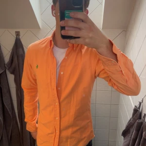 Orange skjorta från Ralph Lauren - Snygg orange skjorta från Polo Ralph Lauren i slim fit-modell. Skjortan har klassisk krage, långa ärmar och knäppning framtill. Broderad grön logga på bröstet ger en clean och fräsch look. Perfekt för dig som vill sticka ut lite extra.