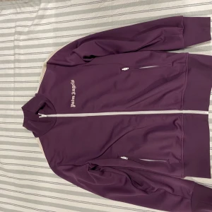 Lila Palm Angels track jacket - Snygg lila track jacket från Palm Angels med vita och svarta ränder längs ärmarna. Jackan har hög krage, hel dragkedja framtill och två fickor med dragkedja. Ikonisk Palm Angels-logga broderad på bröstet. Hör av er vid minsta lilla fundering  pris kan diskuteras vid snabb affär