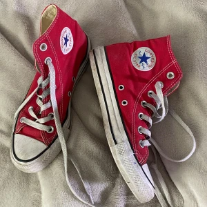 Röda Converse Chuck Taylor All Star - Klassiska röda Converse Chuck Taylor All Star high-tops med vit sula och vita snören. Skorna har rund tå, tydliga logopatchar på sidan och är tillverkade i canvas. Perfekta för en cool och avslappnad stil.