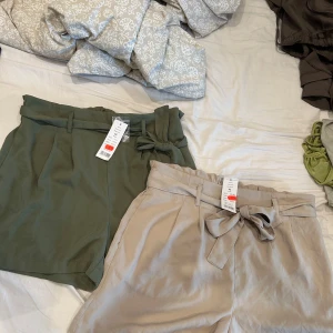 Beige och gröna shorts med knyt - Två par snygga shorts från Gina, ett par i beige och ett i olivgrönt. Båda har hög midja, bälte med knyt och avslappnad passform. Perfekta för varma dagar och lätta att matcha med olika toppar. Materialet känns mjukt och svalt mot huden. 40kr styck