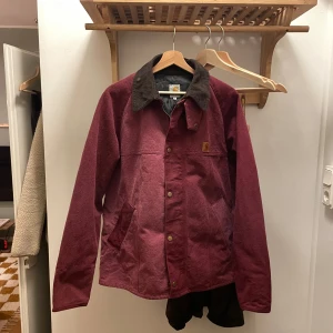Carhartt jacka - Snygg vinröd jacka från Carhartt med kontrasterande mörk krage i manchester. Jackan har knappar framtill, två snedställda fickor och klassisk Carhartt-logga på bröstet. Perfekt för dig som gillar streetwear och vill ha en stilren look. Storleken är XXL men jag skulle säga motsvarar svensk L.