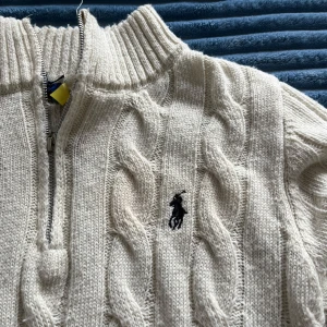 Vit kabelstickad tröja Polo Ralph Lauren - Vit kabelstickad tröja från Polo Ralph Lauren med halv dragkedja och hög krage. Klassisk broderad logga på bröstet. Tjockt, mysigt material i ullblandning och långa ärmar. Perfekt för lager-på-lager under kyliga dagar.