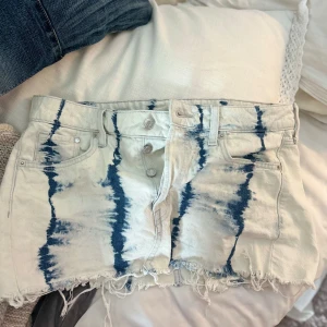 Vit och blå tie-dye jeanskjol - Snygg kort jeanskjol från IDENIM i vit och blå tie-dye med rå, fransig kant. Kjolen har klassiska jeansfickor, bälteshällor och knappar framtill. Perfekt för dig som vill sticka ut med en cool och avslappnad stil.