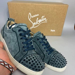 Blå mocka sneakers från Louboutin - Unika blå sneakers i mocka från Christian Louboutin med coola svarta nitar på tån och klassisk röd sula. Skorna har mörkblå snörning och vit platt sula. Storlek 39 men passar som 40