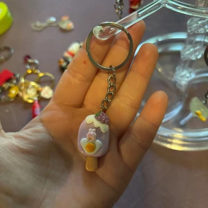 Lila glassformad nyckelring med dekoration - Supergullig nyckelring med en glassformad berlock i lila och vitt, dekorerad med små detaljer som en kanin, ägg och bär. Perfekt accessoar för att piffa upp väskan eller nyckelknippan.