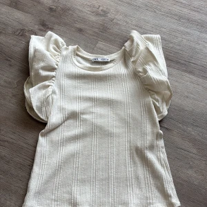 Vit ribbad topp med volangärm Zara - Superfin vit ribbad topp från Zara med breda volangärmar som ger en extra fin touch. Toppen är ärmlös med rund halsringning och har en mjuk, stretchig känsla. Perfekt att styla med jeans eller kjol för en trendig look.