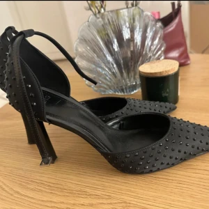 Svarta pumps med nitar och spetsig tå - Snygga svarta pumps med spetsig tå och hög klack. Skorna är täckta av små svarta nitar för en edgy look och har en elegant knytning runt vristen. Materialet är matt och känns lyxigt, perfekt för att sticka ut.