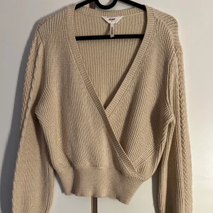 Beige omlott tröja från Object - Superfin beige stickad tröja från Object med omlott-design och djup v-ringning. Tröjan har ribbade muddar, långärmade ärmar med snyggt flätmönster längs sidorna och en croppad passform. Perfekt att slänga på sig när det är kyligt ute!