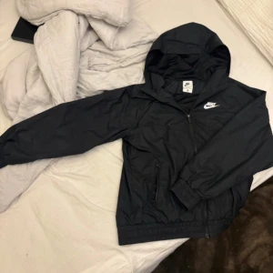 Svart Nike vindjacka med huva - Svart vindjacka från Nike med klassisk logga på bröstet. Jackan har huva, dragkedja framtill och elastiska muddar vid ärmslut och midja. Perfekt för blåsiga dagar och enkel att matcha med streetwear. Tillverkad i lätt polyester för en sportig look.