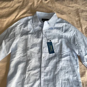 Ljusblå skjorta från Polo Ralph Lauren - Klassisk ljusblå skjorta från Polo Ralph Lauren i bomull. Skjortan har lång ärm, knappar framtill och broderad logga på bröstet. Snygg krage och klassisk passform som funkar till många olika stilar. Perfekt för dig som gillar stilrena och tidlösa plagg. Ingen retur.