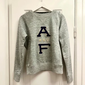 Grå sweatshirt från Abercrombie & Fitch - Snygg grå sweatshirt från Abercrombie & Fitch med stora bokstäver 'A&F' i mörkblått och gult på framsidan. Tröjan har långa ärmar och ribbade muddar vid ärmslut. Nypris ca 700kr