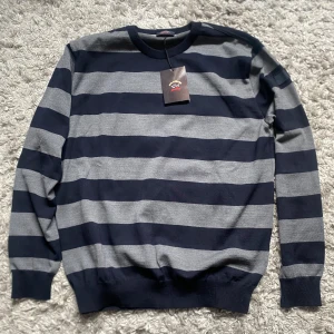Randig tröja från Paul & Shark - Snygg långärmad tröja från Paul & Shark med breda marinblå och grå ränder. Klassisk rund halsringning och ribbade muddar. Perfekt för dig som gillar stilren design med sportig känsla. Materialet är mjuk bomull som känns skönt mot huden. Den är helt ny och aldrig använd, säljs pga att den inte passade mig och inte går att lämna tillbaka längre. Nypris är runt 3000. Hör av er vid fler frågor!