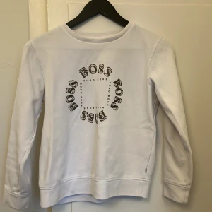 Vit sweatshirt från Hugo Boss - Vit sweatshirt från Hugo Boss med rund hals och tryck i svart på bröstet där det står 'BOSS' i cirkel. Tröjan har långa ärmar och en klassisk, avslappnad passform. Perfekt för en clean och stilren look.