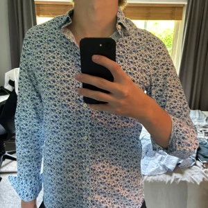 Blommig skjorta från GANT, vit/blå - Säljer en fitted skjorta från GANT i storlek S med blommigt mönster i blått på vit botten. Skjortan har långa ärmar, klassisk krage och knäppning framtill. Tillverkad i bomull, Hör av dig vid frågor eller funderingar!😁