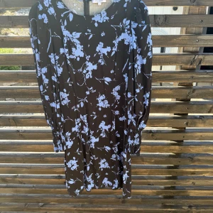 Svart blommig klänning Claire - Svart klänning från Claire med ljusblått blommönster. Modellen är rak och har långa ärmar med smock vid handlederna. Klänningen slutar ovanför knäna och har en mjuk, flowy känsla. Perfekt för dig som gillar en feminin och trendig stil.