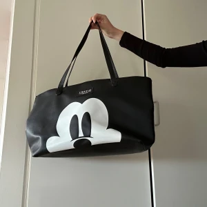 Svart shoppingväska Mickey Mouse - Stor svart shoppingväska från Disney med ett stort vitt tryck av Mickey Mouse ansikte på framsidan. Väskan har långa handtag och är gjord i syntetmaterial, perfekt för att bära allt du behöver. Snygg och lekfull design som sticker ut.