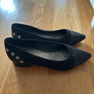 Svarta ballerinaskor med nitar från Micha - Vi kan komma överens om priset! Svarta ballerinaskor från Micha med spetsig tå och låg klack. Skorna har en cool ormskinnsstruktur på tå och häl, samt silvriga nitar baktill för extra edge. Materialet är mocka och skinnimitation, perfekt för dig som gillar detaljer.