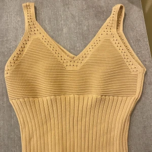 Beige ribbad linnetopp med håldetaljer - Snygg beige linnetopp med ribbad struktur och smala axelband. Toppen har v-ringning och dekorativa håldetaljer längs ringningen. Materialet är stretchigt och sitter tight mot kroppen, perfekt för en trendig och enkel look.