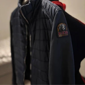 Parajumpers cardigan (BYTEN) - Säljer en mörkblå quiltad jacka från Parajumpers med emblem på ärmen. Jackan har dragkedja framtill och är långärmad med släta ärmar och quiltad kropp. Perfekt för dig som vill ha en stilren och sportig look.