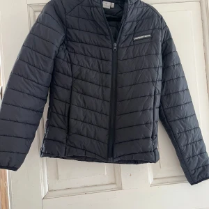 Svart lättviktsjacka från Cross Sportswear - Svart quiltad lättviktsjacka från Cross Sportswear med dragkedja framtill och diskret logga på bröstet. Jackan är tillverkad i nylon och har en klassisk passform med långa ärmar. Perfekt för lager-på-lager under kyliga dagar. Har ett stygn som gått upp på väster sida. Annars i super fint skick. 