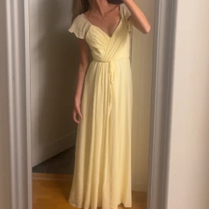 Ljusgul långklänning från Bubbleroom - Superfin ljusgul långklänning från Bubbleroom med omlottliv, volangdetaljer vid axlarna och öppen rygg med knyt. Perfekt för sommarens alla tillfällen, men köpt för att användas på bal för 2 årsen. Använd en gång. 