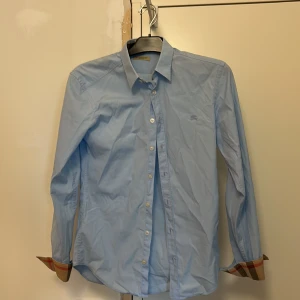 Ljusblå skjorta från Burberry - Säljer en klassisk ljusblå skjorta från Burberry Brit i storlek XS. Skjortan har långa ärmar, knappar framtill och den ikoniska Burberry-rutiga detaljen på insidan av manschetterna. Perfekt för en stilren look.