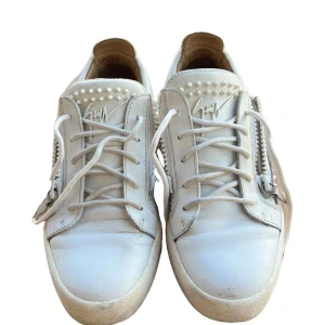 Vita sneakers med nitar och dragkedja från Giuseppe Zanotti - Säljer ett par vita sneakers från Giuseppe Zanotti med coola nitar på häl och plös. Skorna har dragkedjor på sidorna och snörning framtill. Tillverkade i skinn med rund tå och platt sula. Perfekta för dig som gillar detaljer och streetstyle.