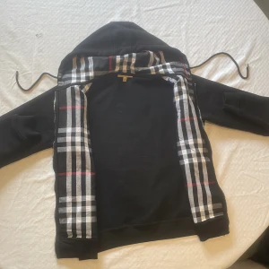 Svart hoodie med dragkedja från Burberry - Säljer en svart hoodie från Burberry med dragkedja och klassiskt rutigt foder i huvan. Jackan har broderad logga på bröstet och snörning i huvan. Perfekt för en stilren och avslappnad look.