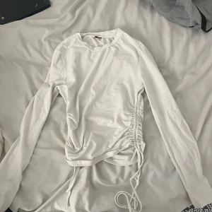 Vit långärmad topp med snörning - Säljer en vit långärmad topp med trendig snörningsdetalj på sidan som ger en snygg rynkad effekt. Toppen har rund halsringning och är i mjukt bomullsmaterial. Perfekt för en stilren och modern look.