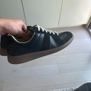 liknande Maison Margiela skor - Säljer ett par klassiska svarta sneakers som är en exakt liknande modell som märket Maison Margiela med vita snören och bruna gummisulor. Skorna har en mix av mocka och skinn på ovandelen och en rund tå. Perfekta för dig som gillar stilrena och tidlösa sneakers.
