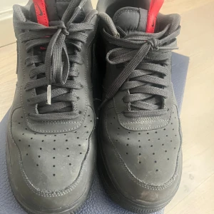 Nike Air Force 1 i svart med röda detaljer - Säljer ett par svarta Nike Air Force 1 med röda detaljer på plösen och hälen. Skorna har klassisk rund tå, snörning och är tillverkade i skinn. Perfekta för dig som gillar stilrena sneakers med en twist.