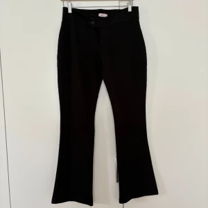 Svarta bootcut byxor med låg midja  - Säljer ett par svarta byxor med bootcut-modell och låg midja. Byxorna har två knappar framtill och är i ett stretchigt material som sitter snyggt över höfterna. Perfekta till både vardag och när du vill känna dig extra snygg.