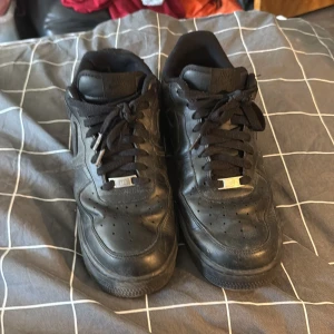 Svarta Nike Air Force 1 sneakers - Klassiska helsvarta Nike Air Force 1 sneakers med svart snörning och rund tå. Skorna har en platt sula och är tillverkade i skinn. Perfekta för en stilren och enkel look.