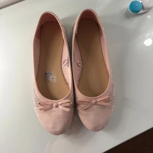 Ljusrosa ballerinaskor med rosett och strass - Säljer ett par ljusrosa ballerinaskor med dekorativ rosett framtill och små glittrande strass på sidorna. Skorna har rund tå och platt sula, perfekta för en söt och feminin look.