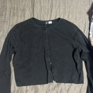 Svart croppad kofta från Divided - Svart croppad kofta från Divided med rund halsringning och knäppning framtill. Långärmad och i mjukt bomullsmaterial, perfekt att styla över ett linne eller t-shirt.