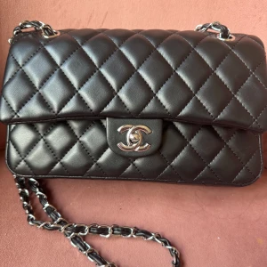 Helt ny! Svart quiltad double flap axelväska i lambskin - Säljer en klassisk svart axelväska i lambskin i quiltat skinn med silverfärgad kedja och ikonisk märke framtill. Insidan är vinröd och har präglad skrift inne i väskan som det ska. Perfekt för dig som vill ha en tidlös och lyxig accessoar. Inte använd. För stor för mig därför säljer jag denna grymt fina kvalité  på materialet och snygga väskan. Jag har lagt det priset tyvärr tycker många men det är otrolig kvalitékommer inte sänka pris heller utan  behåller själv då. Vill ni köpa så gör det. 😊