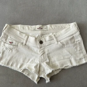 Vita jeansshorts från Hollister - Säljer ett par vita jeansshorts från Hollister med råa kanter och klassisk femficksmodell. Shortsen har låg midja och en liten röd logga på fickan framtill. Perfekta för varma dagar!