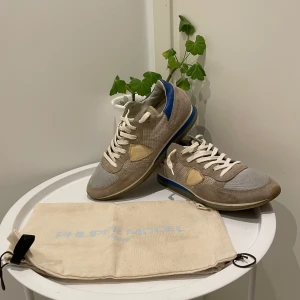 Philippe Model sneakers i beige och blå - Säljer ett par Philippe Model sneakers i beige mocka och mesh med vita snören och blå detalj vid hälen. Skorna har rund tå och platt sula. Perfekta för en avslappnad och stilren look.