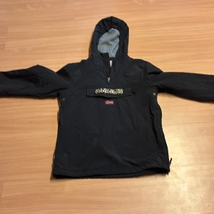 Svart vindjacka med huva från Napapijri - Svart vindjacka från Napapijri med huva och halv dragkedja framtill. Jackan har en stor ficka på magen med norsk flagga och logotyp. Priset är inte hugget i sten så bara att höra av er vid funderingar 