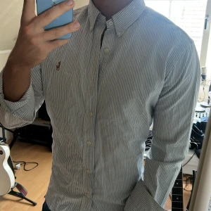 Blårandig skjorta från Ralph Lauren - Klassisk blårandig skjorta från Ralph Lauren med button-down krage och broderad logga på bröstet. Skjortan har långa ärmar och vita knappar. Perfekt för en stilren och tidlös look.