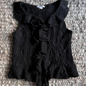 Svart volangblus från Gina Tricot - Säljer en svart ärmlös blus från Gina Tricot med volangdetaljer längs kragen och framtill. Blusen har en lätt skrynklig struktur och en feminin, elegant look. Perfekt att styla till olika outfits.