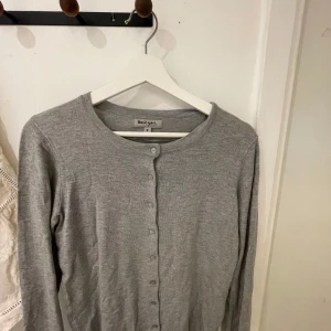 Grå kofta från Basic You - En enkel och stilren grå kofta från Basic You i storlek S. Koftan har rund halsringning, långa ärmar och knäppning framtill med små knappar. Perfekt att slänga över en topp eller klänning för en avslappnad look.