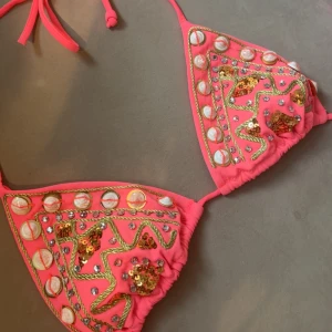 Bikinitop - Superfin trekantsbikini i starkt rosa/lite orange med guldiga paljetter, snäckor och glittriga stenar. Bikinin har knyt i nacken och ryggen samt dekorativa broderier och snörning. SJUKT SNYGG. Säljer då den tyvärr är för liten. 