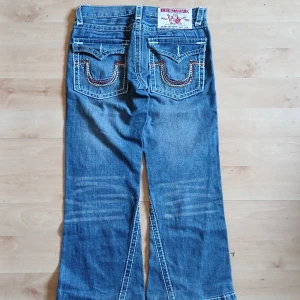 Blå jeansbyxor med vita sömmar från True Religion - Säljer ett par blå jeansbyxor från True Religion med tydliga vita kontrastsömmar och broderade bakfickor. Klassisk femficksmodell med bootcut och låg midja. Perfekta för dig som gillar en avslappnad men snygg stil.