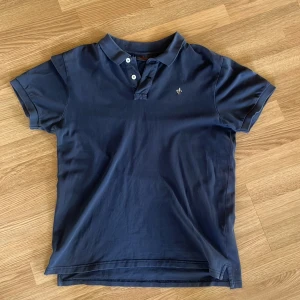 Mörkblå pikétröja från Morris - Snygg mörkblå pikétröja från Morris med klassisk krage och knappar framtill. Tröjan har korta ärmar och en broderad logga på bröstet. Perfekt för en stilren och avslappnad look. Inga defekter 