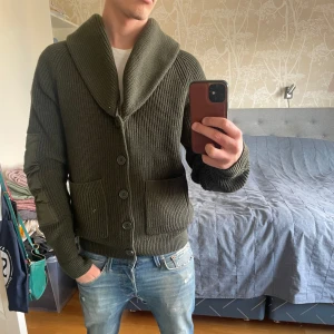 Olivgrön stickad kofta från J.Lindeberg - Säljer en olivgrön stickad kofta från J.Lindeberg med stora knappar framtill, två fickor och bred krage. Ärmarna har snygga quiltade detaljer i samma färg. Perfekt för lager-på-lager och en cool vardagslook. Litet hål nertill på högra armen. 